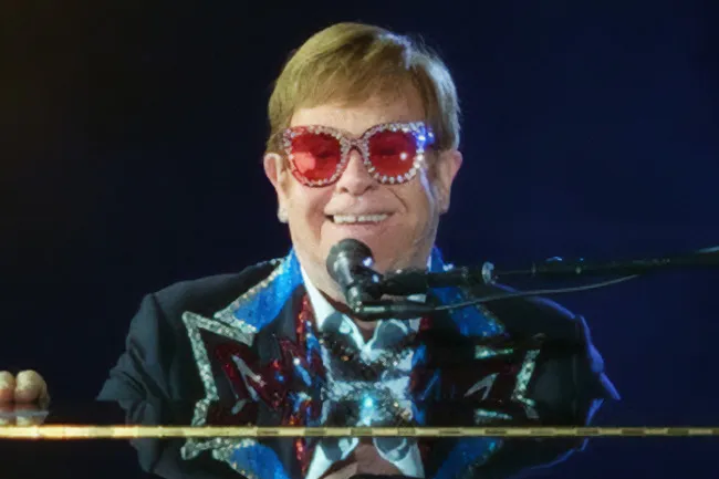 Elton John smiling, wearing large embellished sunglasses and a sequined jacket, playing the piano.