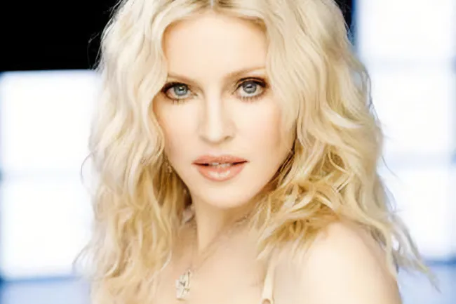 Madonna, a music icon, with long wavy blonde hair, light makeup, and a necklace, looking directly at the camera.