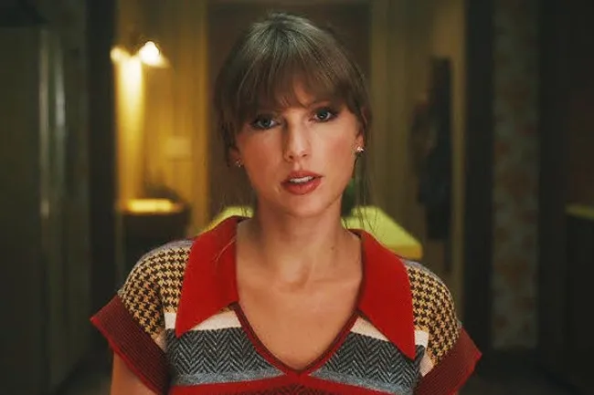 Taylor Swift, with bangs, wearing a red-collared shirt with a houndstooth pattern, looking directly at the camera.