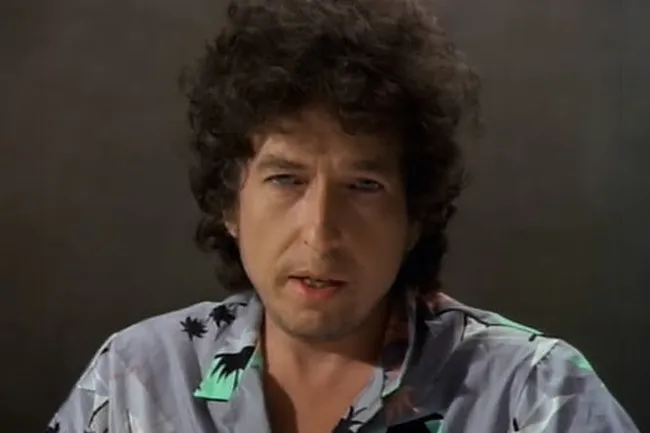Bob Dylan, with curly hair and a mustache, wears a tropical shirt, looking forward with blue eyes. Music icon.