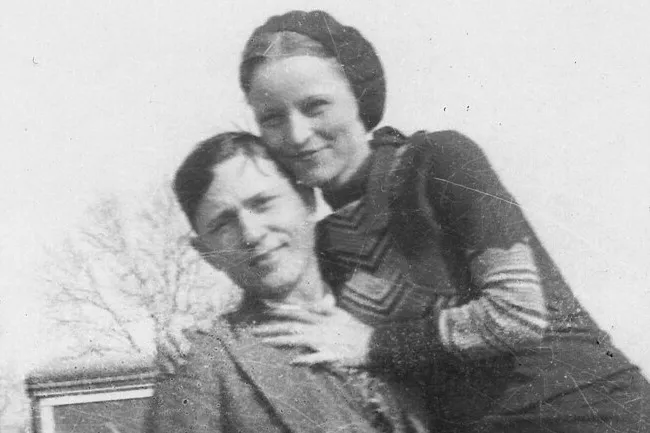 A black and white photo of a young couple smiling, the woman affectionately holds the man. Could they be Bonnie & Clyde?