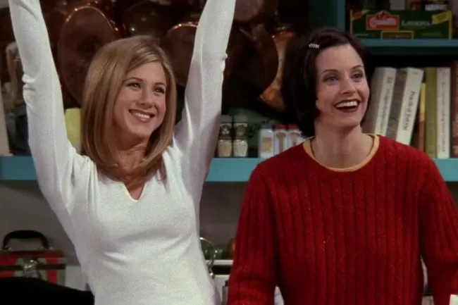 Rachel and Monica, smiling happily. Rachel in a white top with arms raised, Monica in a red sweater.