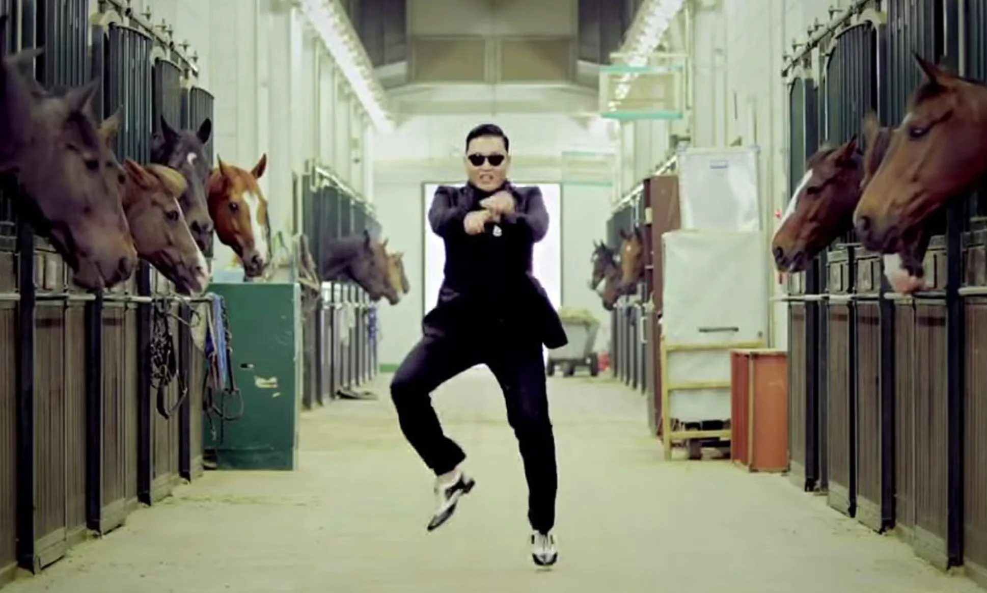Psy in sunglasses and a black suit, doing the Gangnam Style dance in a horse stable, pop culture.