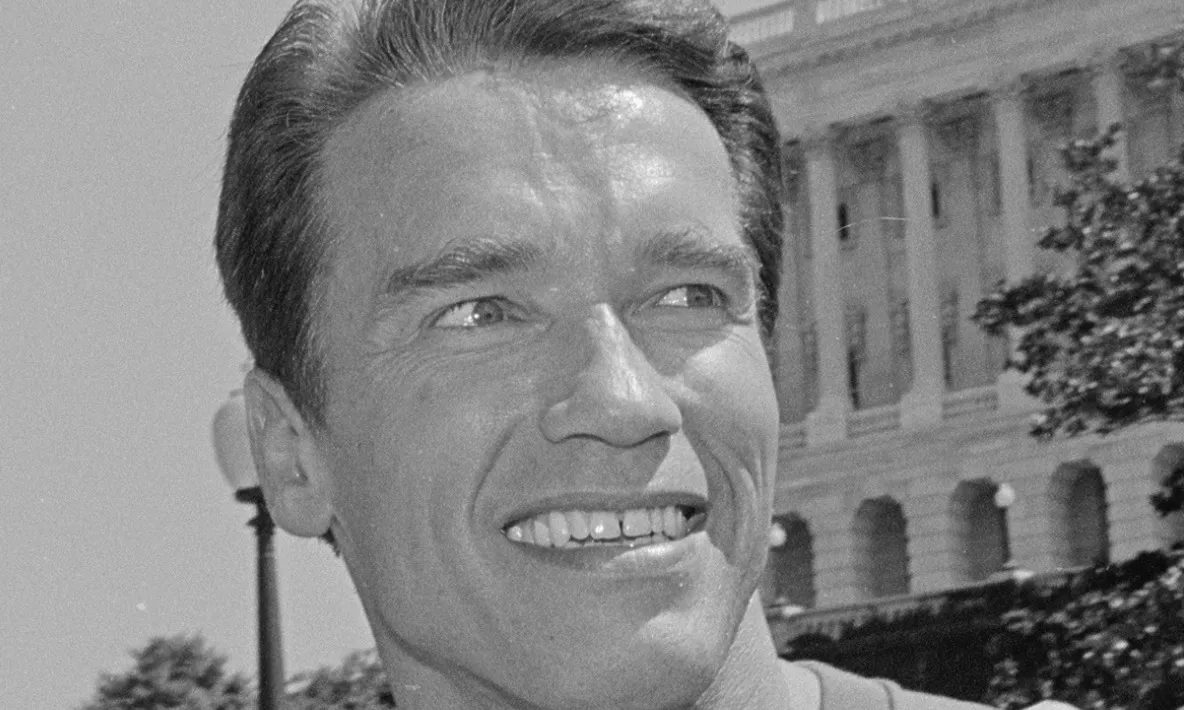 A black and white image of Arnold Schwarzenegger smiling, with a government building in the background. Pop Culture Quiz.