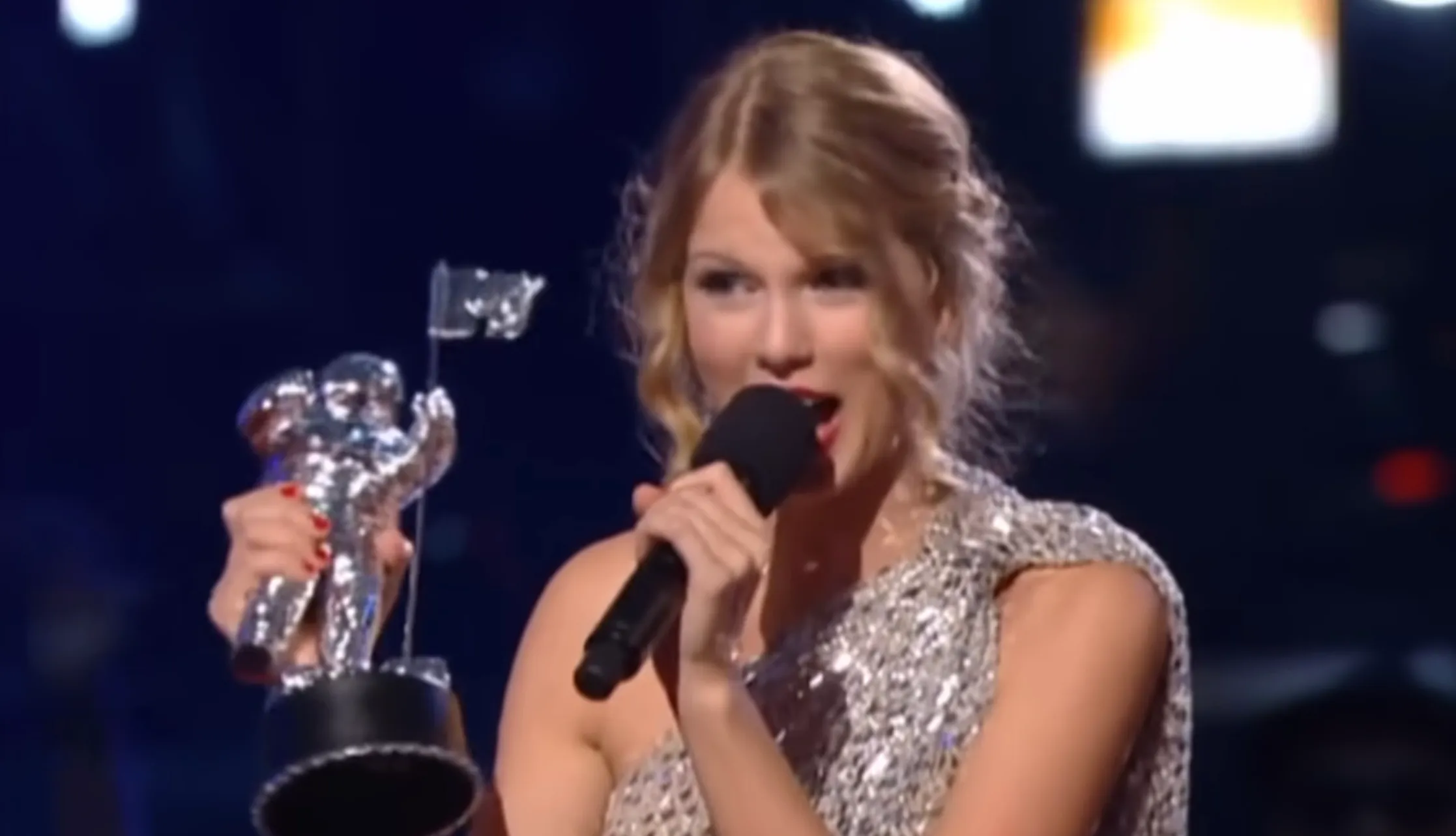 Taylor Swift holds her MTV VMA trophy and microphone, mid-speech. Pop Culture Quiz: Who interrupted Taylor Swift?