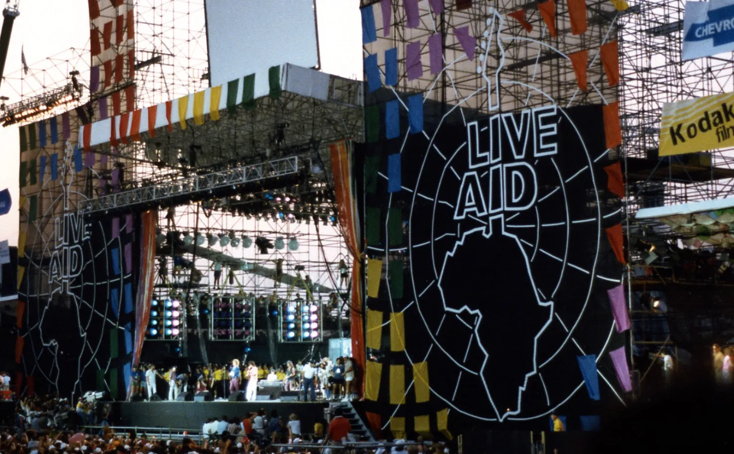 Live Aid stage with performers and massive audience. A legendary pop culture event, featuring a British rock band performance.