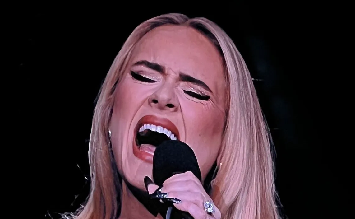 Singer Adele performing live, eyes closed and mouth open, holding a microphone, with a passionate expression.
