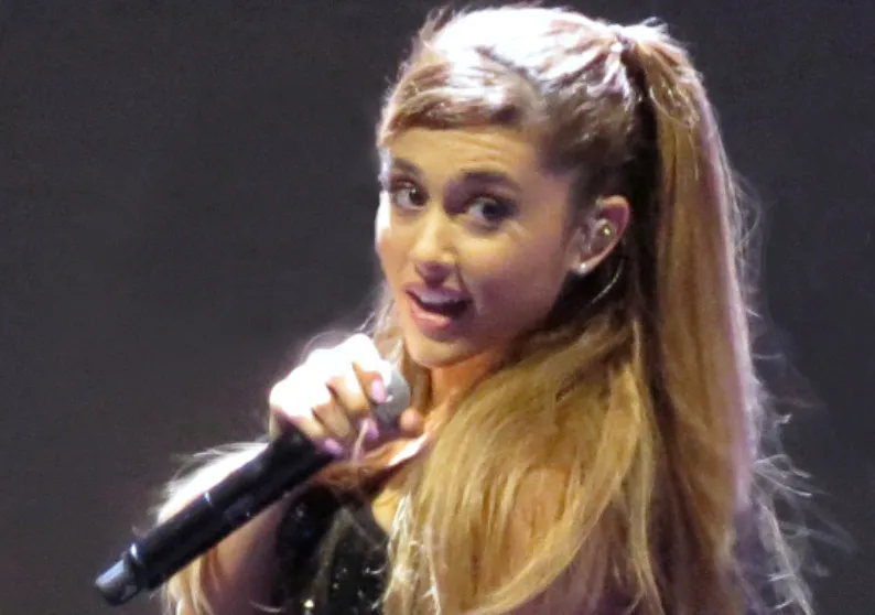 Ariana Grande performing on stage, holding a microphone, with her signature high ponytail and light brown hair.