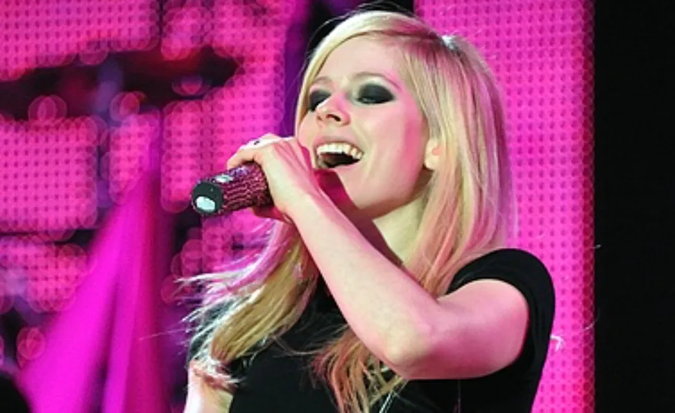 Avril Lavigne performing, holding a microphone, with a bright pink backdrop. She has blonde hair and dark eye makeup.