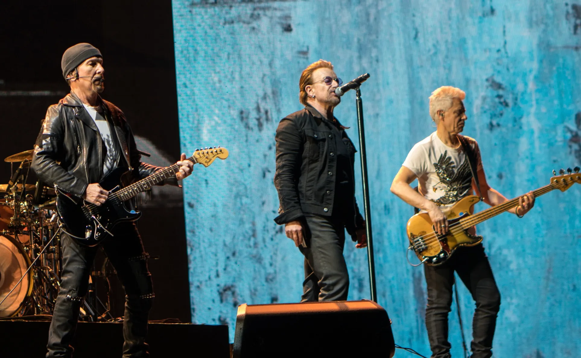 U2 performs live on stage. The Edge plays guitar, Bono sings, and Adam Clayton plays bass.