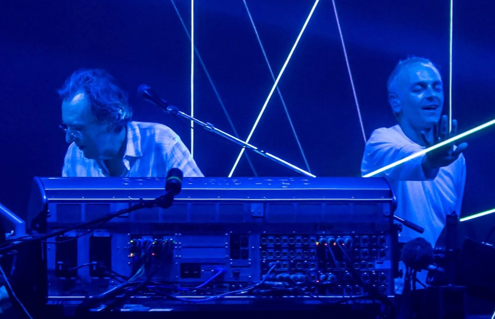Underworld members performing live with synthesizers and blue lighting, glowing lines in the background.