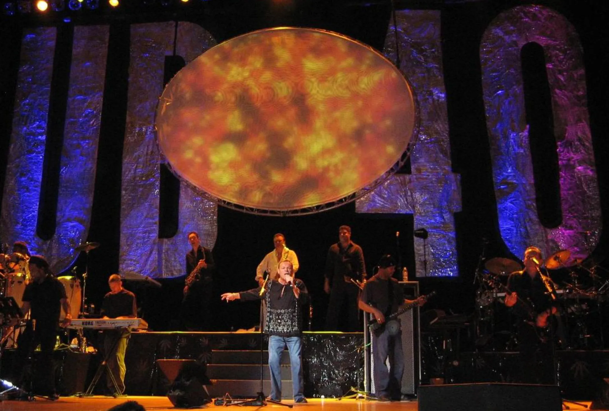 UB40 performs on stage with a large glowing oval screen and the letters "UB40" behind them.