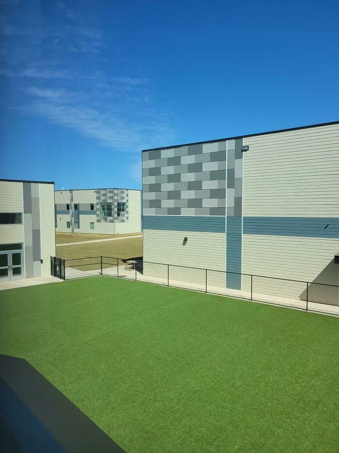 Modern buildings with a large green artificial turf area under a bright blue sky, an unsettling liminal space.