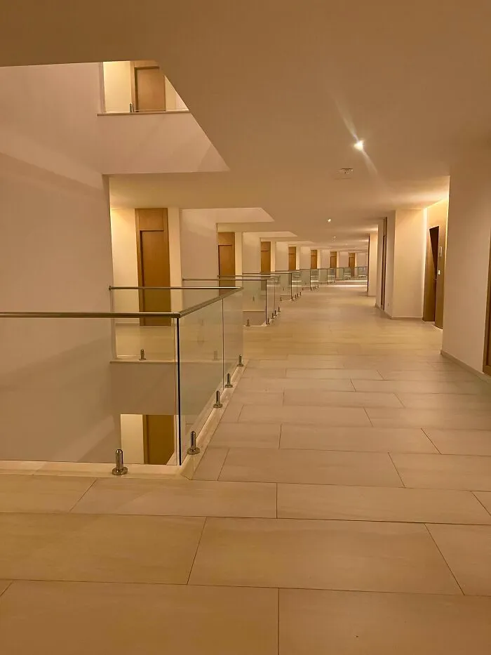 A long, deserted modern hallway with glass railings and many doors, an unsettling liminal space.
