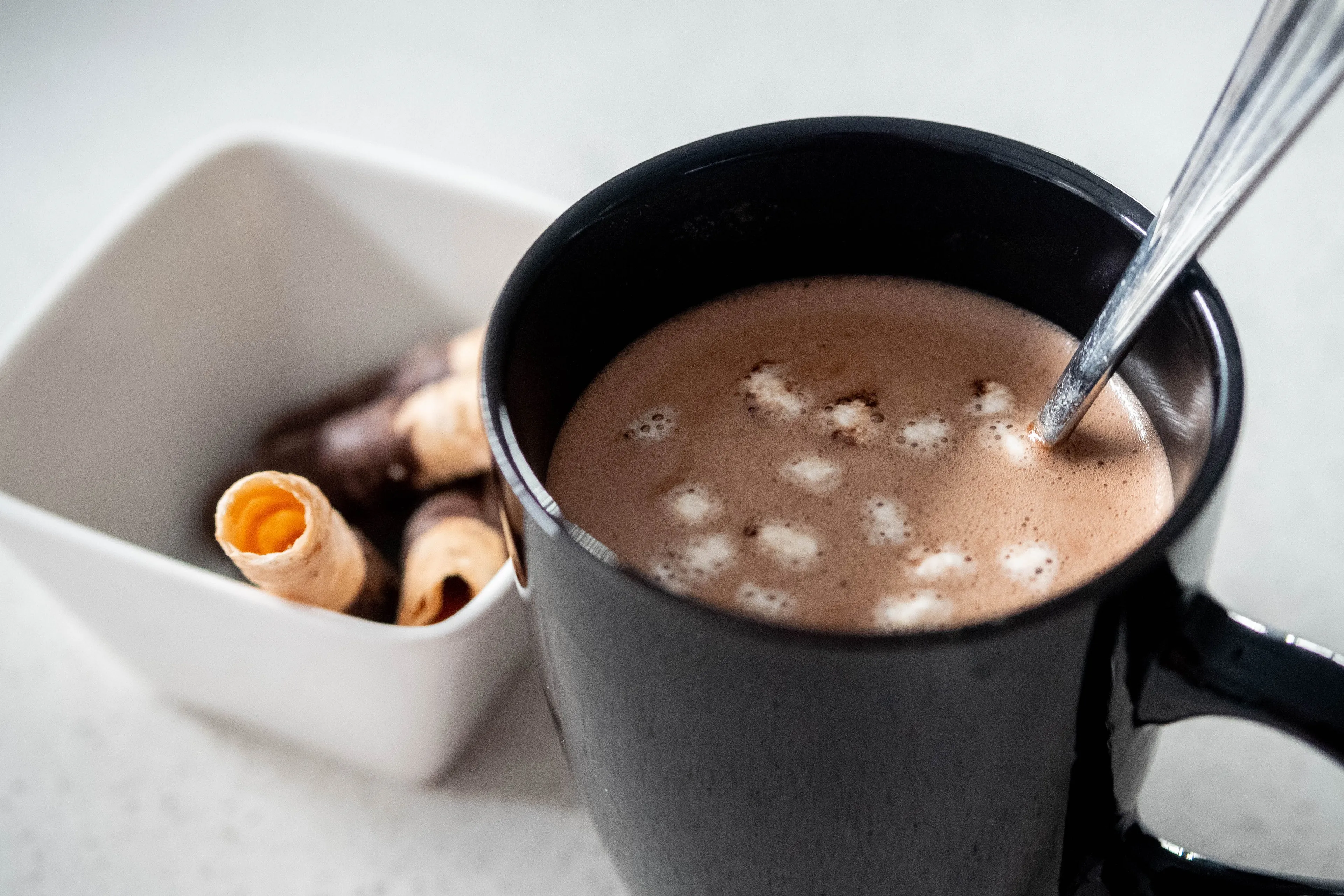 A black mug filled with hot chocolate and mini marshmallows, next to a bowl of waffle rolls, a perfect comfort drink.