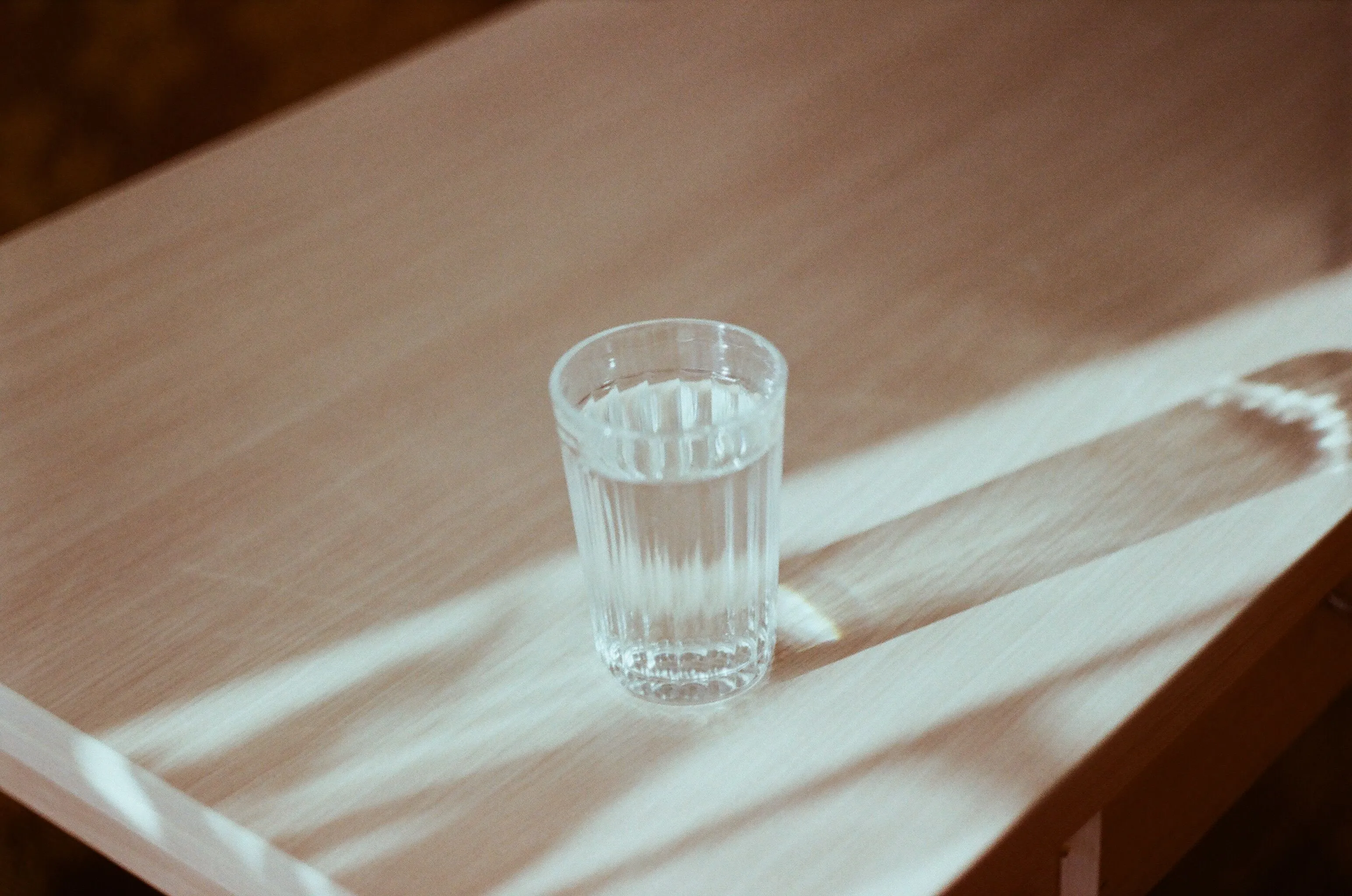 A clear glass of fresh water on a light wooden table with sun streaks, a simple comfort drink.