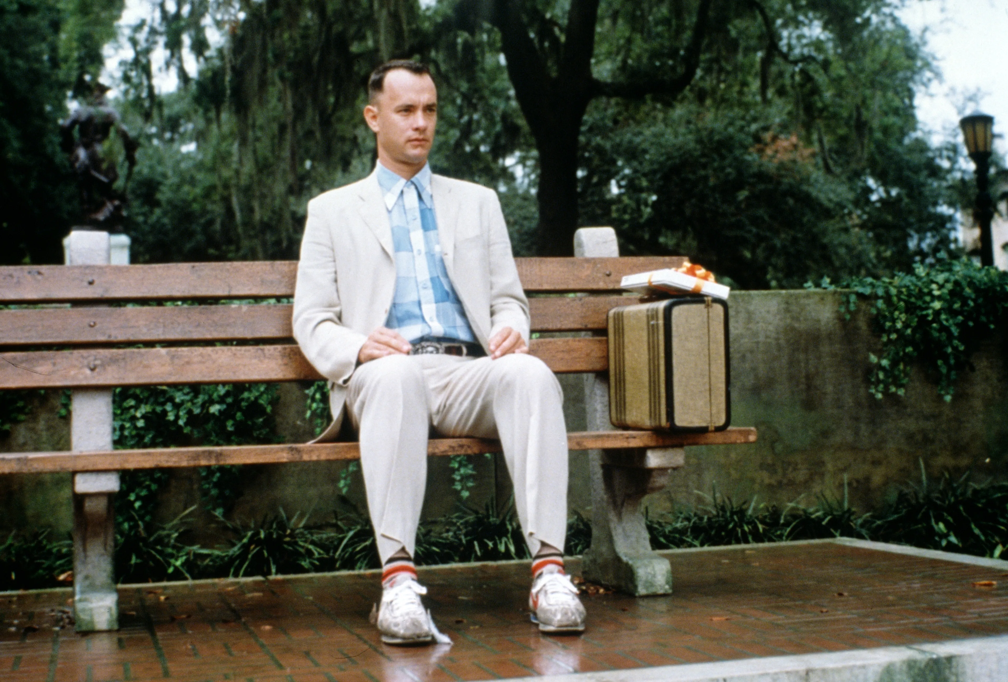 Forrest Gump sits on a park bench with a suitcase and a box, a scene for a movie night alone.