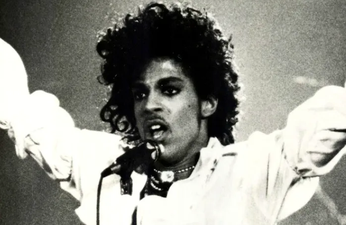 A black and white image of musician Prince singing, with his arms raised, a microphone near his mouth.
