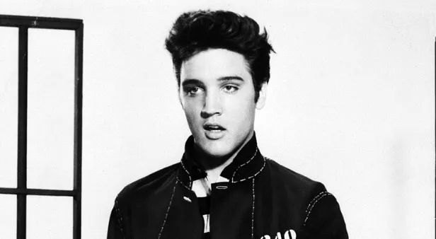 Black and white photo of young Elvis Presley looking up and to the right, wearing a collared jacket. Musician King of Pop.