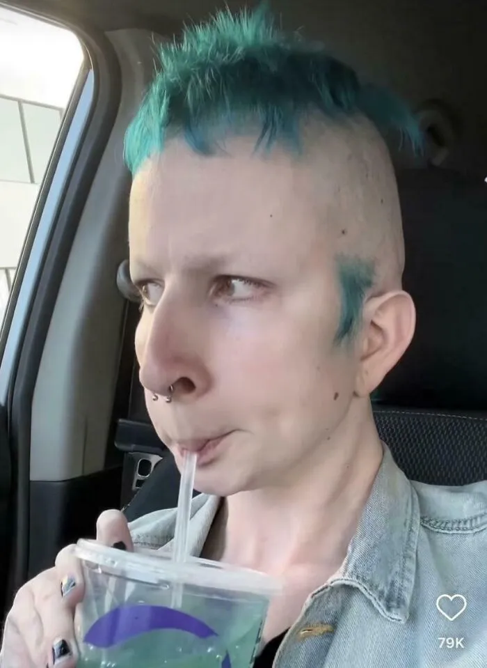 Person with an unfortunate haircut, bright blue hair in a short mohawk, drinking from a cup.