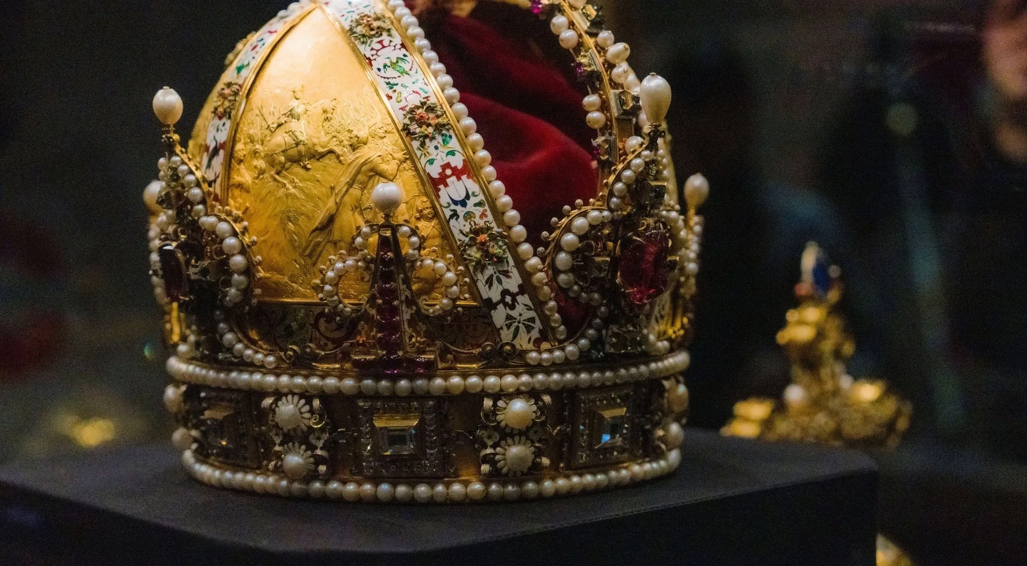 An ornate golden crown, heavily adorned with pearls and gemstones, featuring a red velvet interior. Monarchy in Europe.