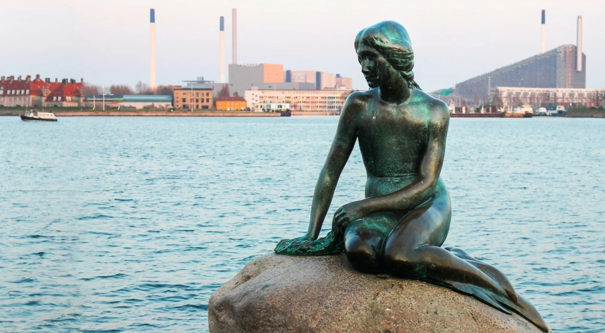 The Little Mermaid statue, a bronze figure with a greenish patina, sits on a rock by the water in Europe.