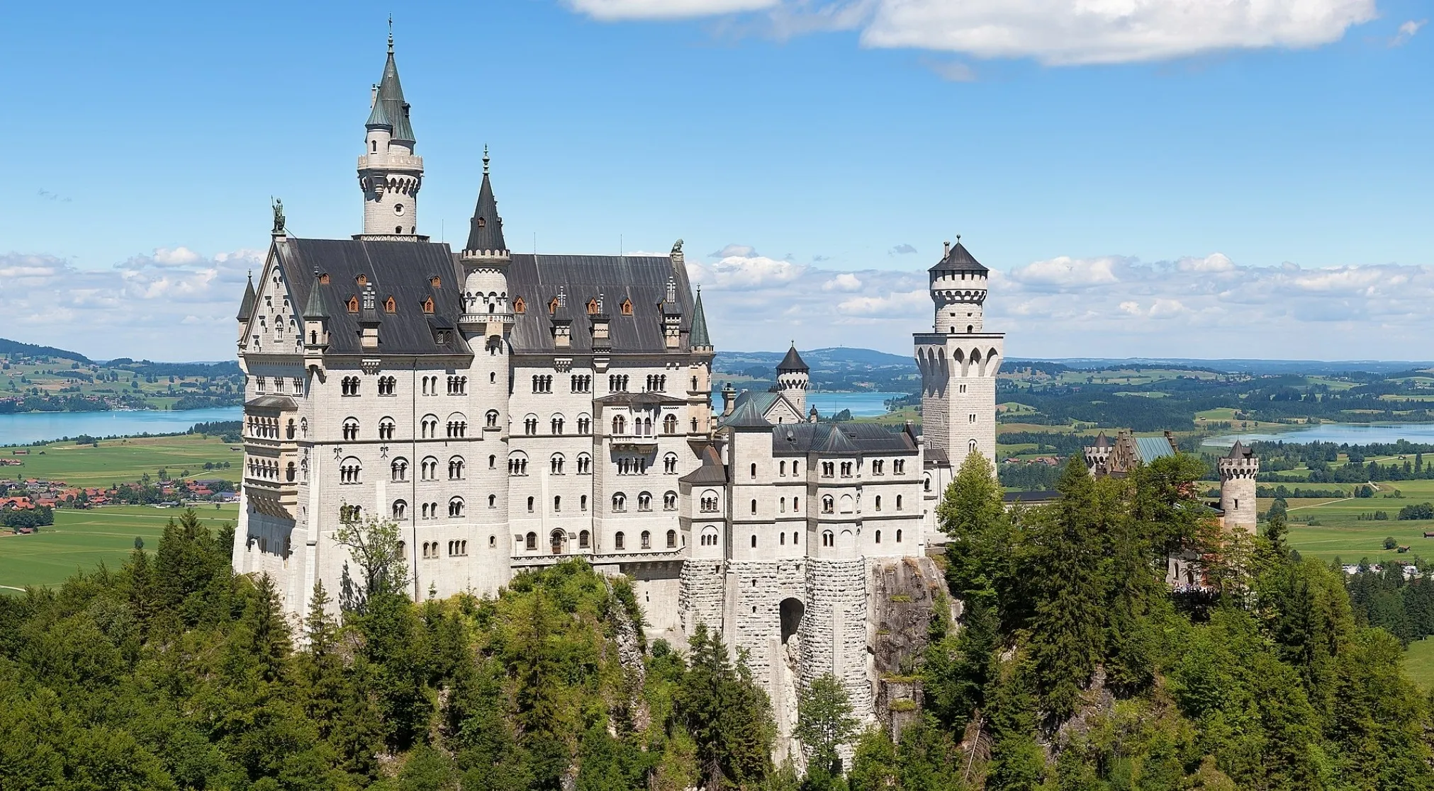Neuschwanstein Castle in Europe, perched on a hill, overlooking lakes and green fields under a blue sky.