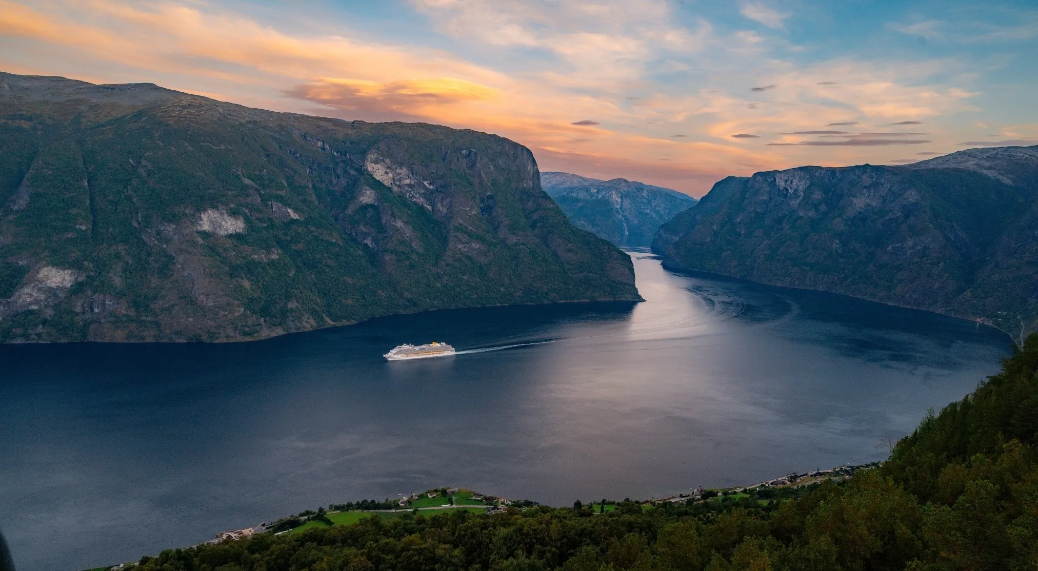 An aerial view of a majestic European fjord with a cruise ship traversing its deep blue waters, surrounded by towering, green mountains under a twilight sky.