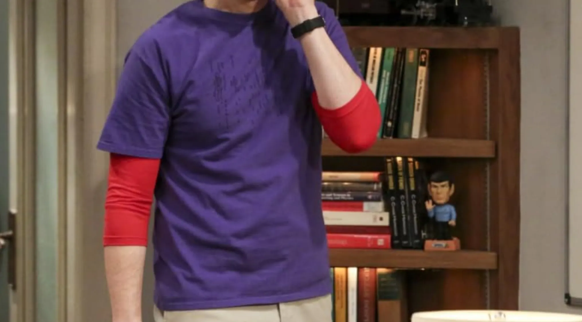 A man wearing a purple t-shirt over a red long-sleeved shirt, standing in a room with a bookshelf, showcasing a recognizable TV show outfit.