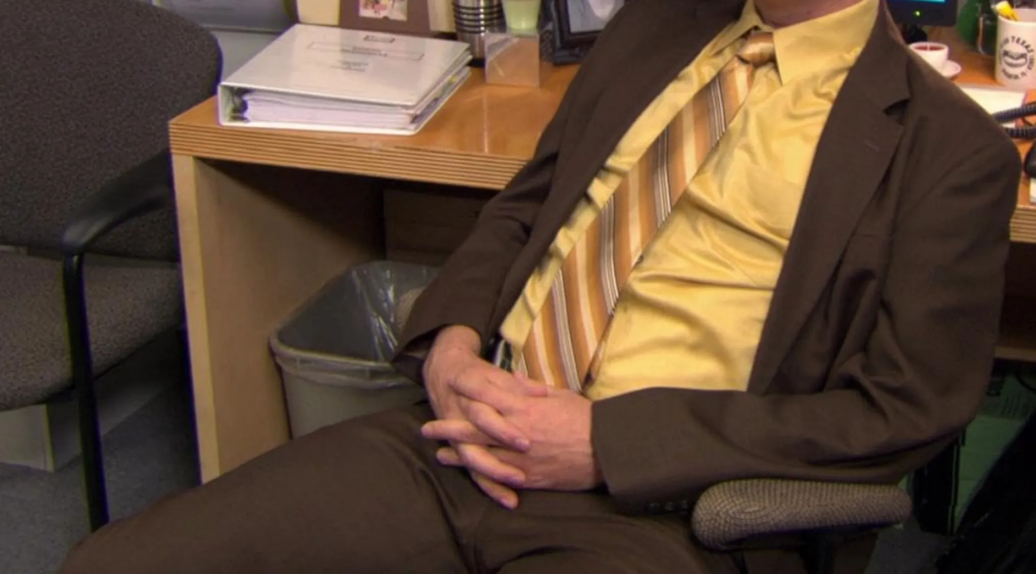 TV Show Outfits: A person seated, wearing a mustard yellow shirt, brown suit, and striped tie.