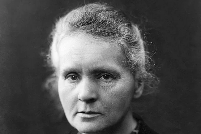A black and white close-up photo of Marie Curie, a historical figure, looking directly at the camera.