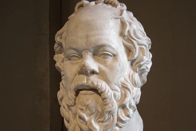 Close-up of a white marble bust of Socrates, showing his bearded face and curly hair against a dark background.