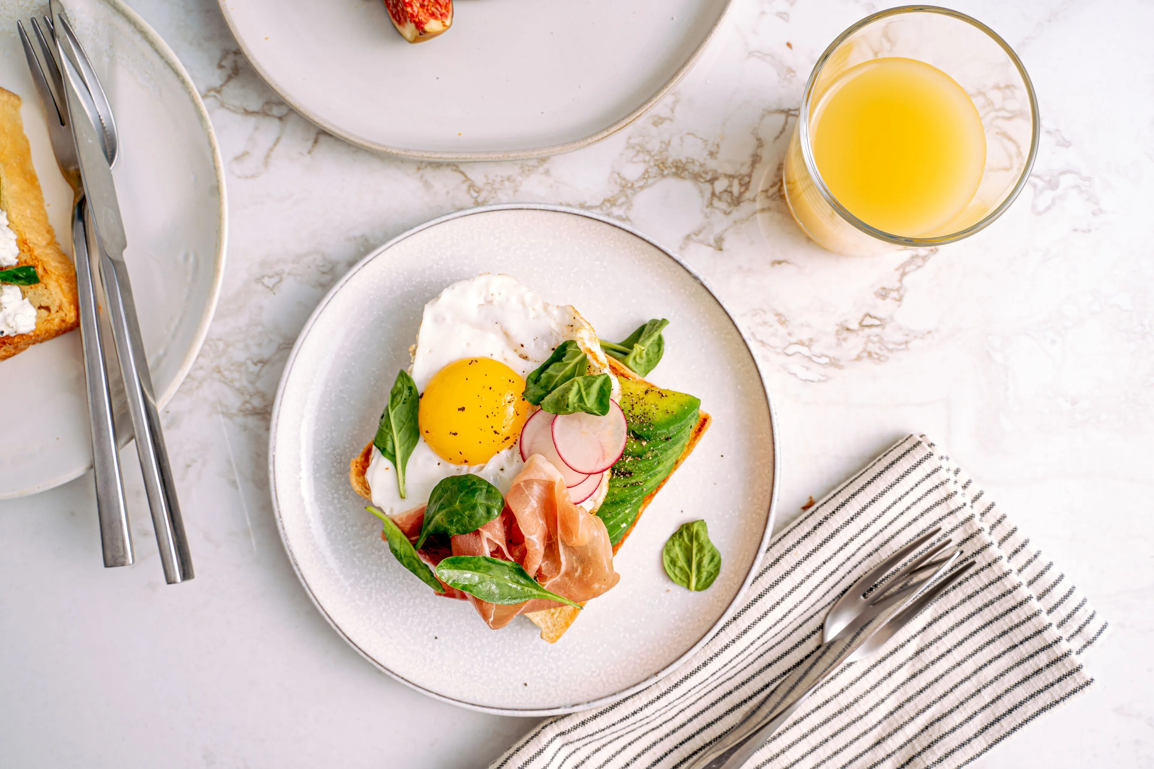 Ideal Saturday morning: making big breakfast with avocado toast, a fried egg, prosciutto, spinach, and radish on a white plate.