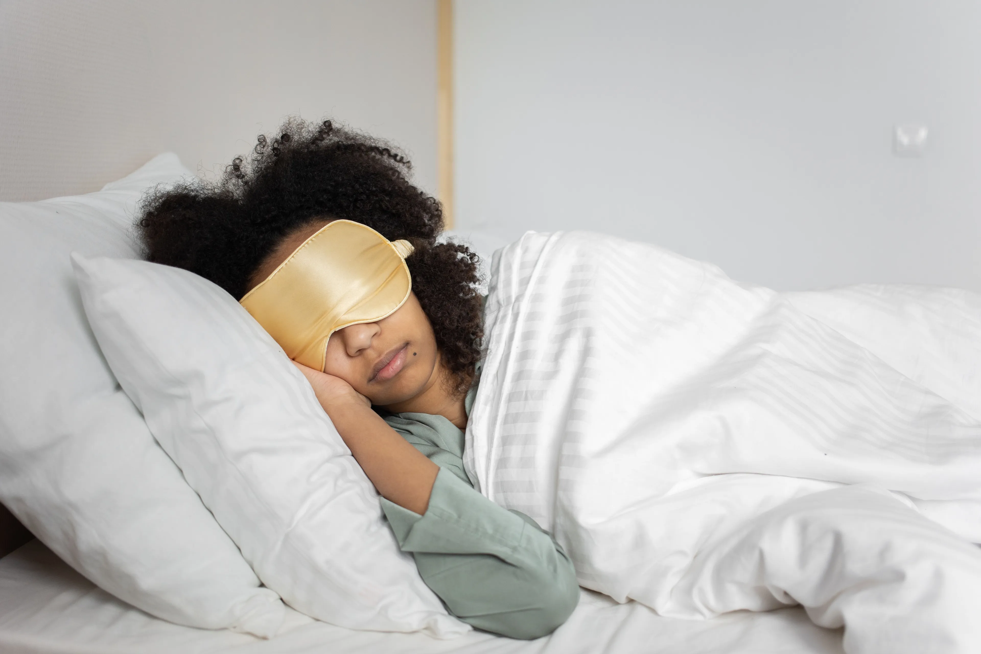 A person with curly hair sleeping in bed, wearing a golden eye mask, enjoying sleeping in on a Saturday morning.