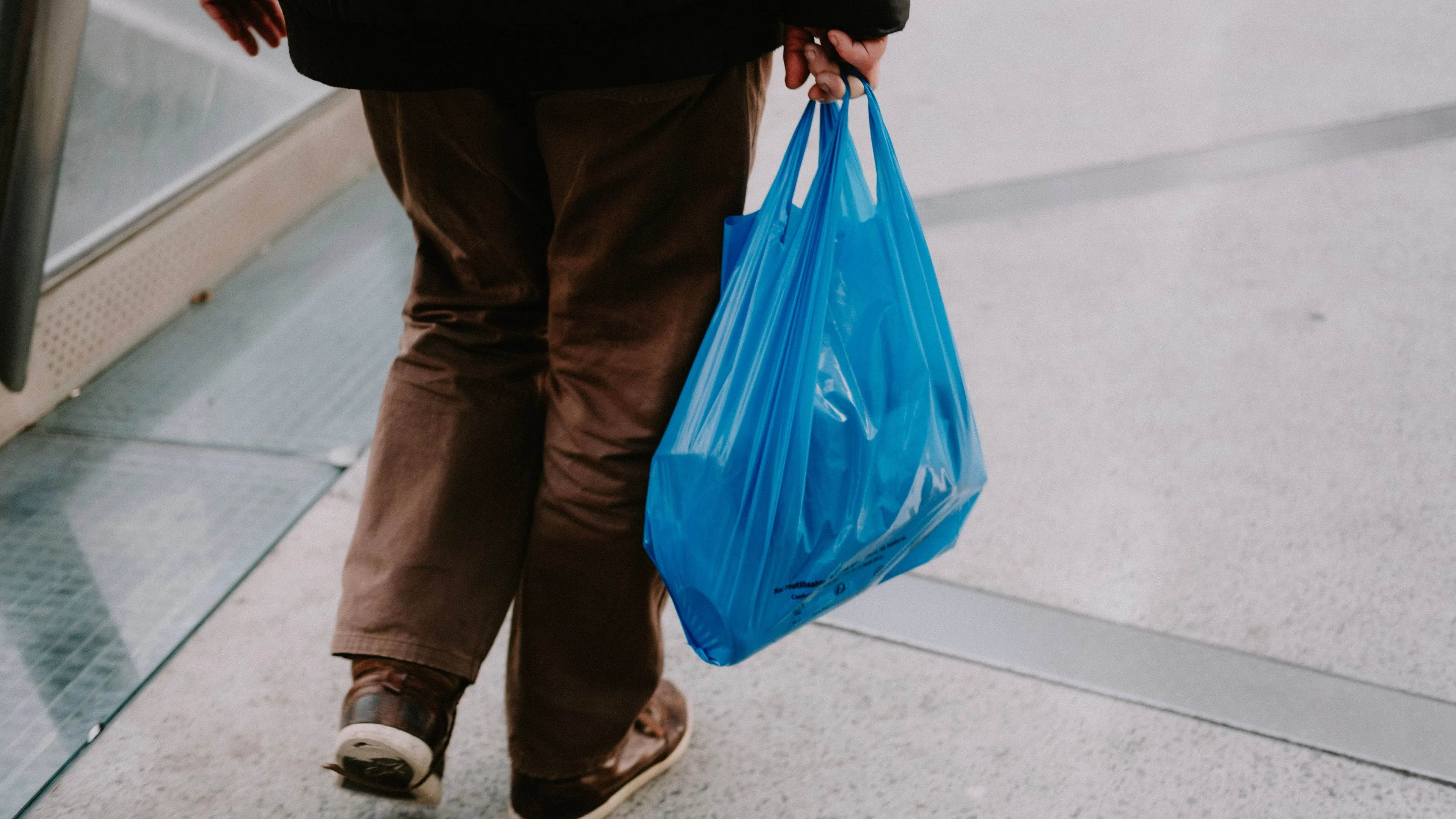 Person in brown pants and shoes, carrying a blue plastic bag, walking outdoors. Perfect for running errands on a Saturday.