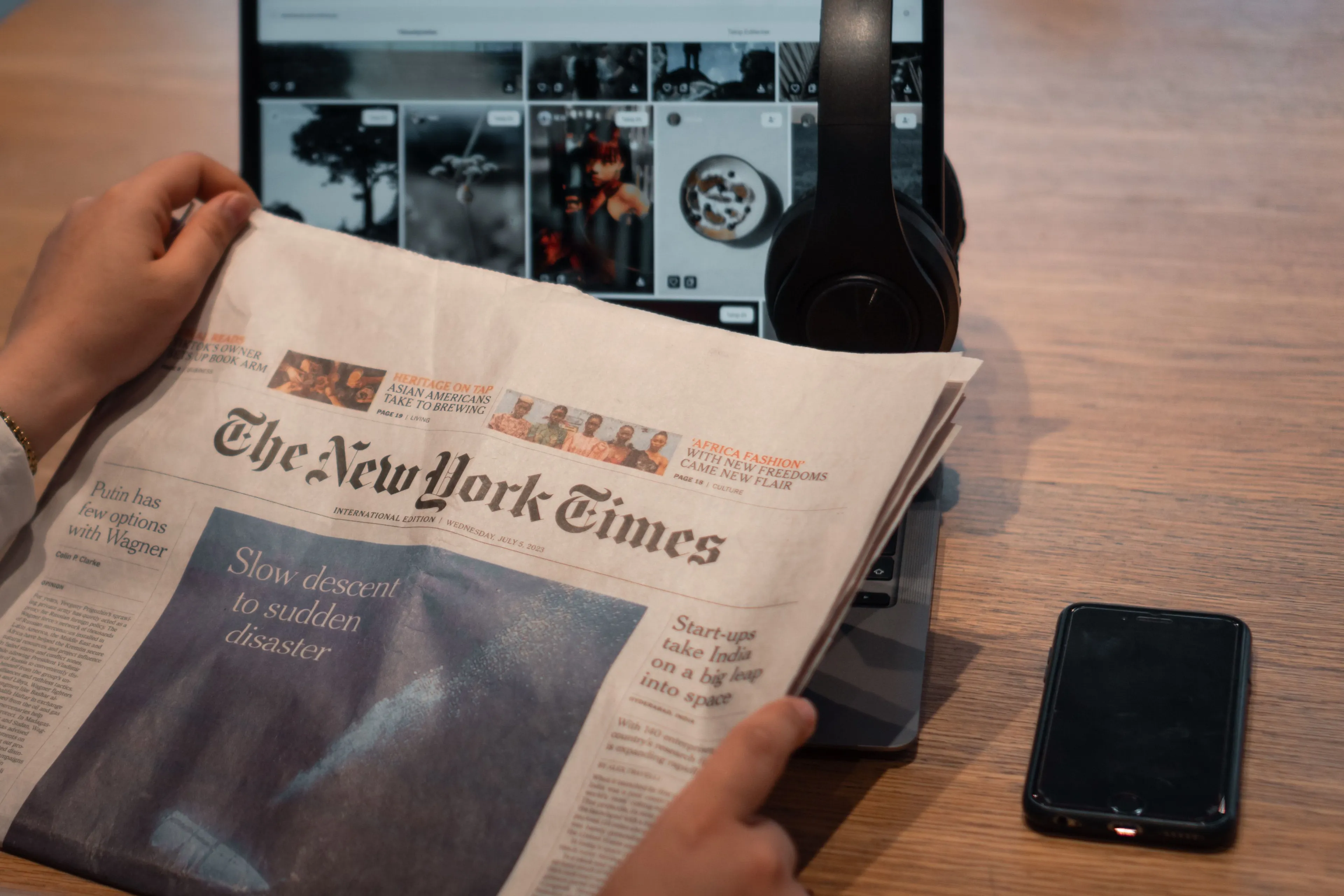 Hands holding The New York Times, reading the news. Laptop with headphones and phone in the background on a wooden desk.