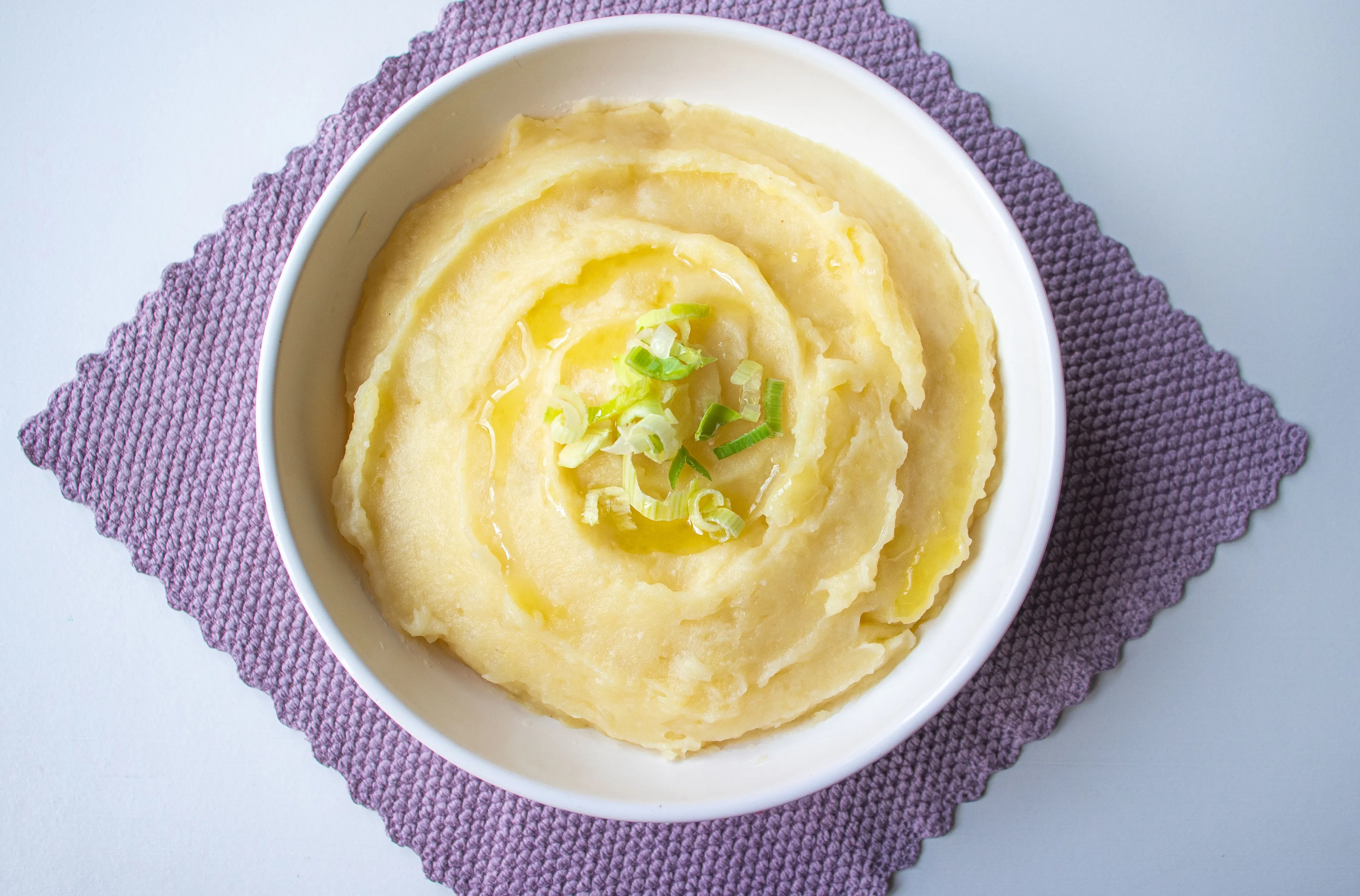 A white bowl filled with creamy mashed potatoes, drizzled with melted butter and garnished with fresh green onions.