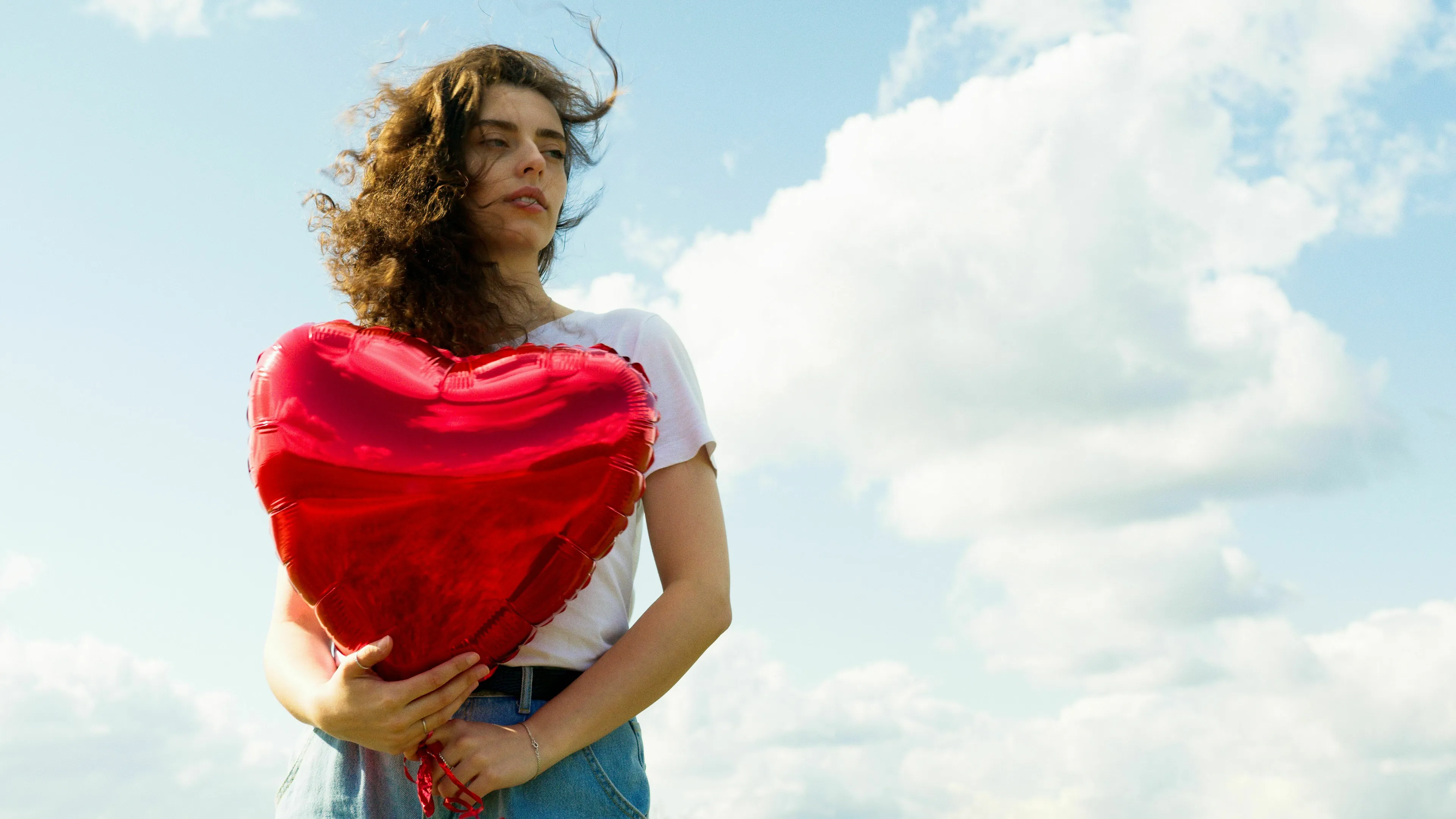 A woman with curly hair holds a red heart balloon under a blue sky, contemplating a red flag or catching feelings.