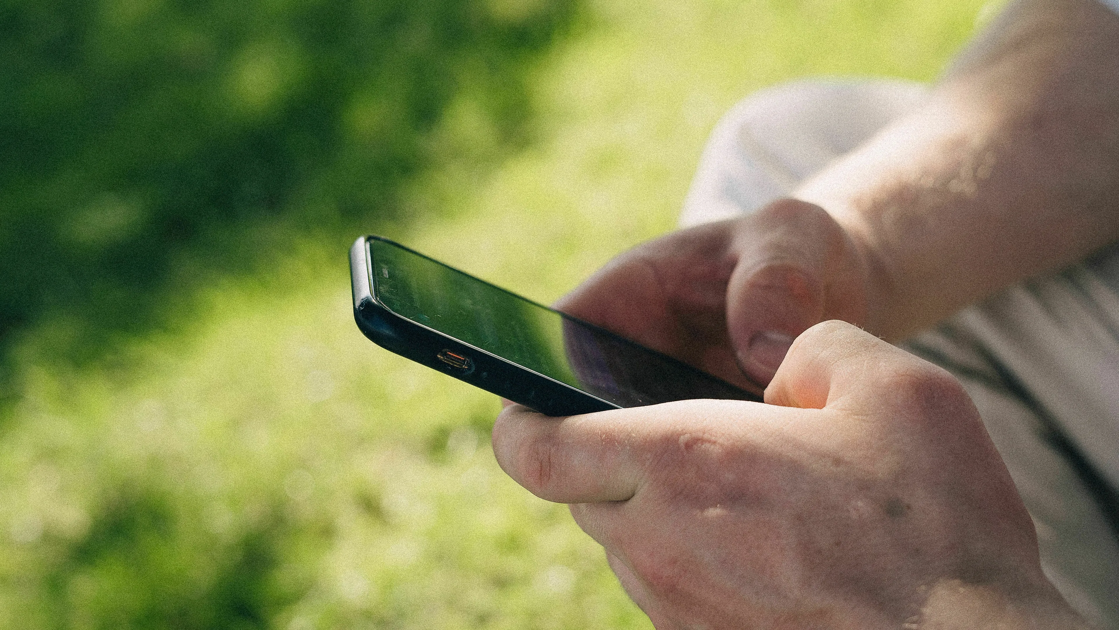 A person's hands holding a smartphone outdoors with a blurred green background. What's your relationship with your phone?