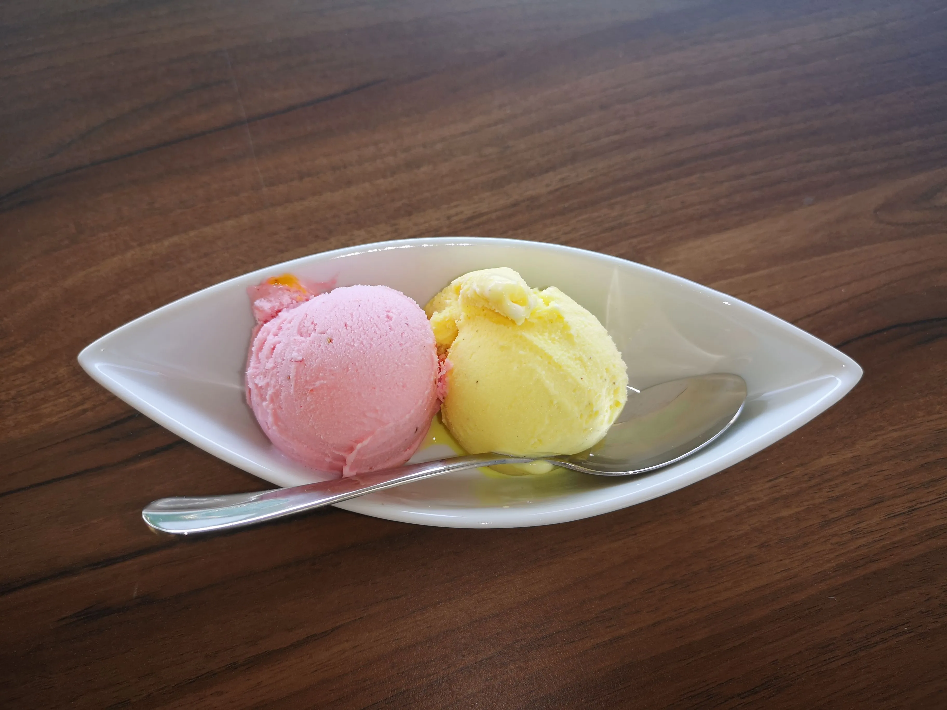 Two scoops of ice cream, one pink and one yellow, in a white bowl with a spoon on a wooden table.
