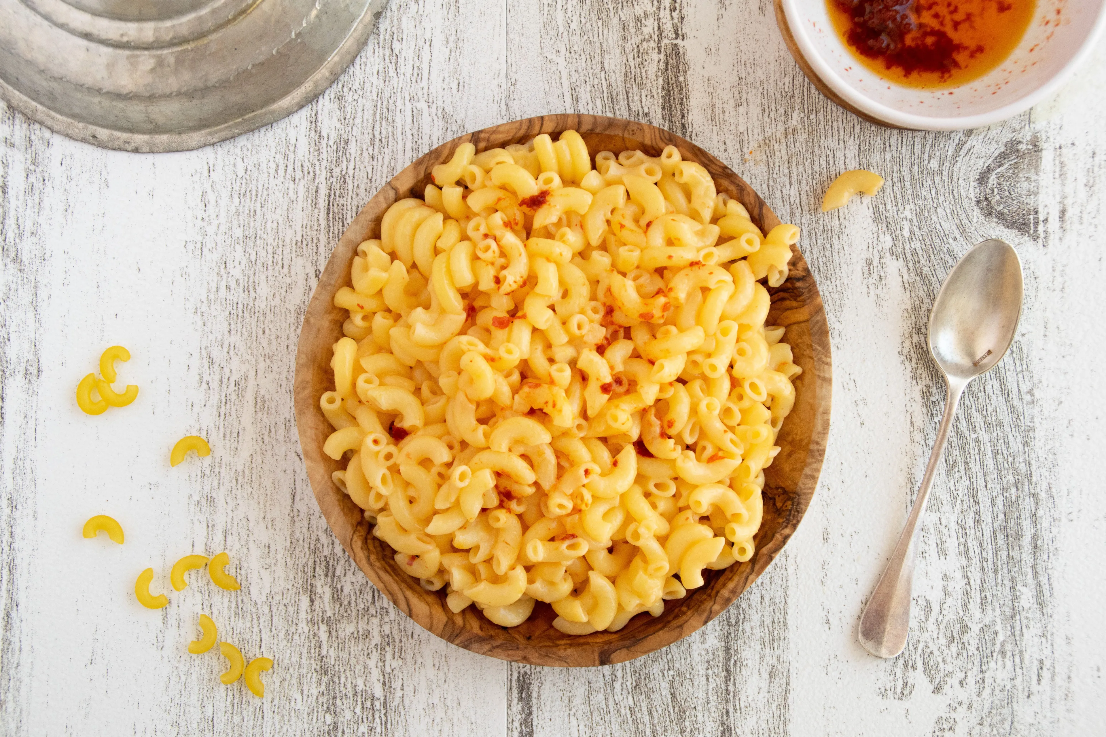 A wooden bowl of creamy mac and cheese, sprinkled with red flakes, on a white wooden table with a spoon and dipping sauce.