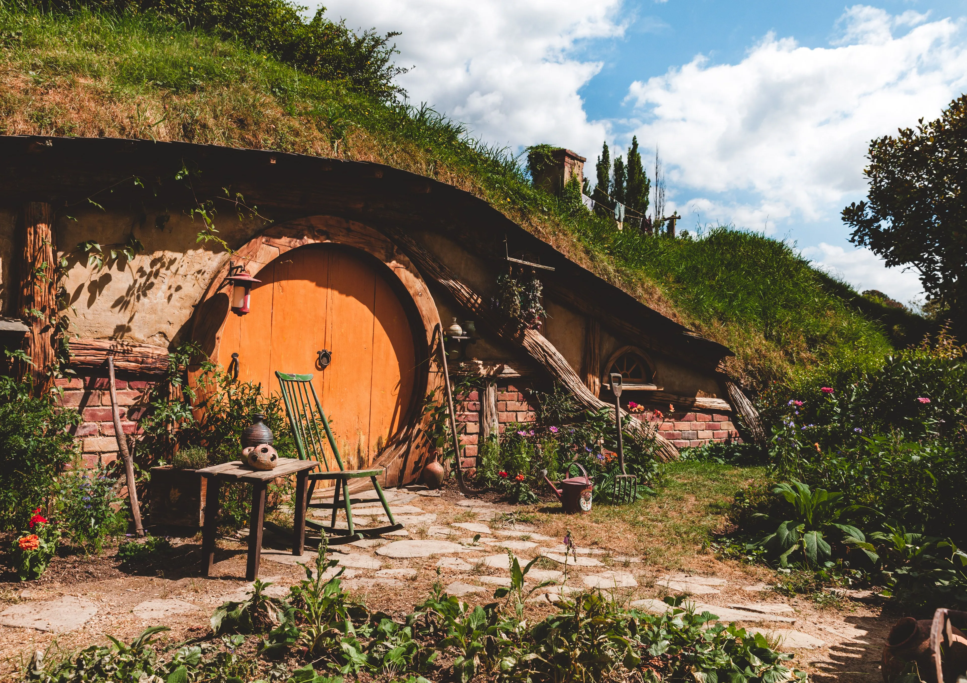 A hobbit-hole in The Shire, with an orange round door, green rocker, and plants, under a blue sky.