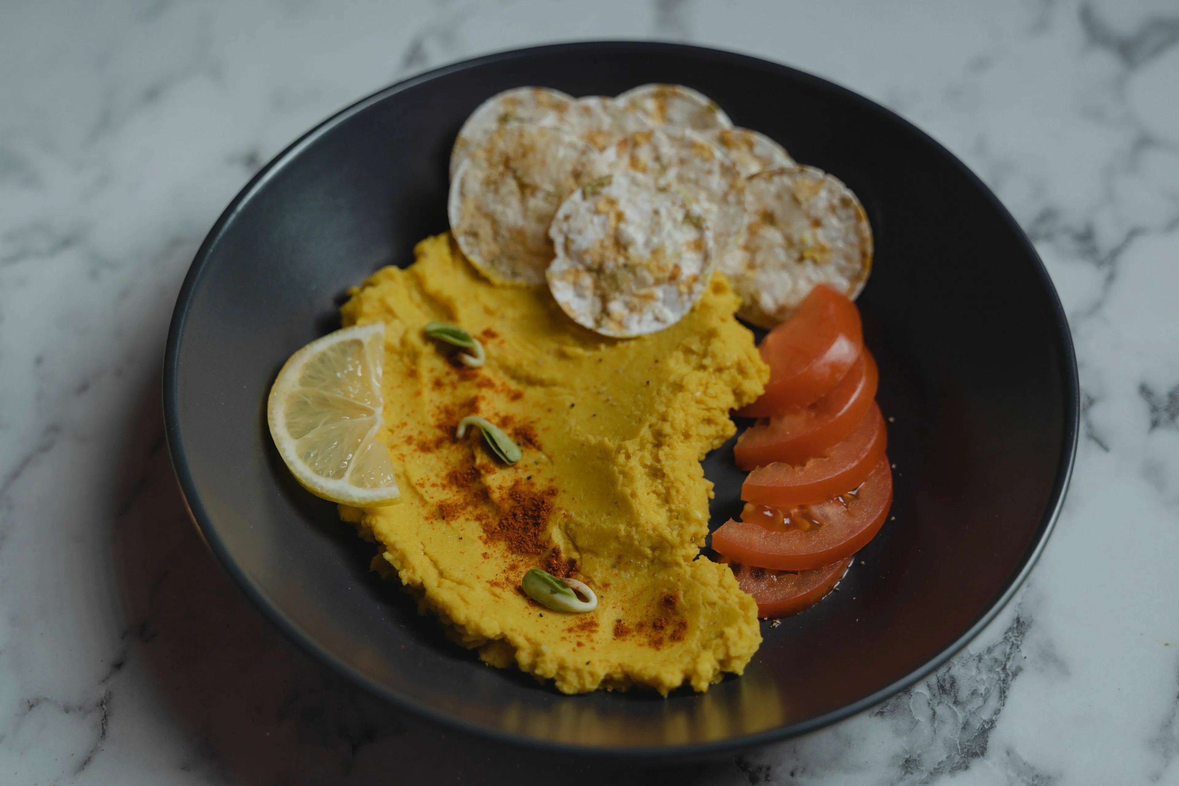 A black plate of yellow hummus, topped with paprika and pistachios, served with rice crackers, tomato slices, and a lemon wedge.