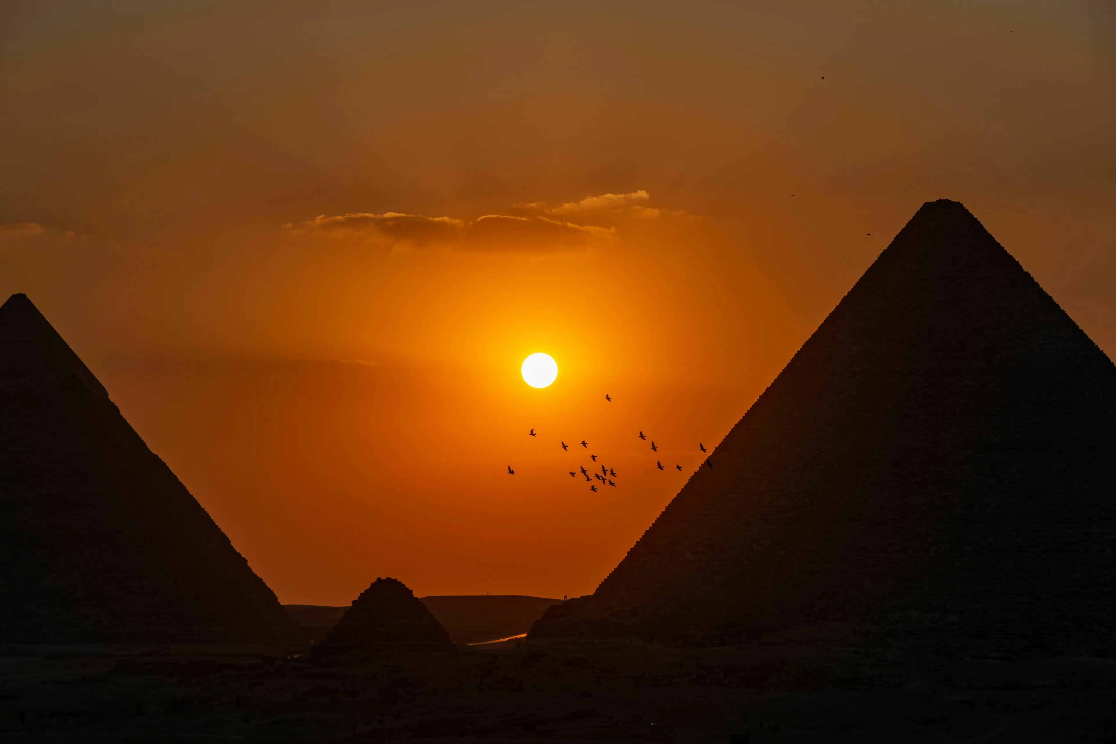 Silhouetted pyramids at sunset with a flock of birds flying past the bright sun god. Egyptian mythology, general knowledge.
