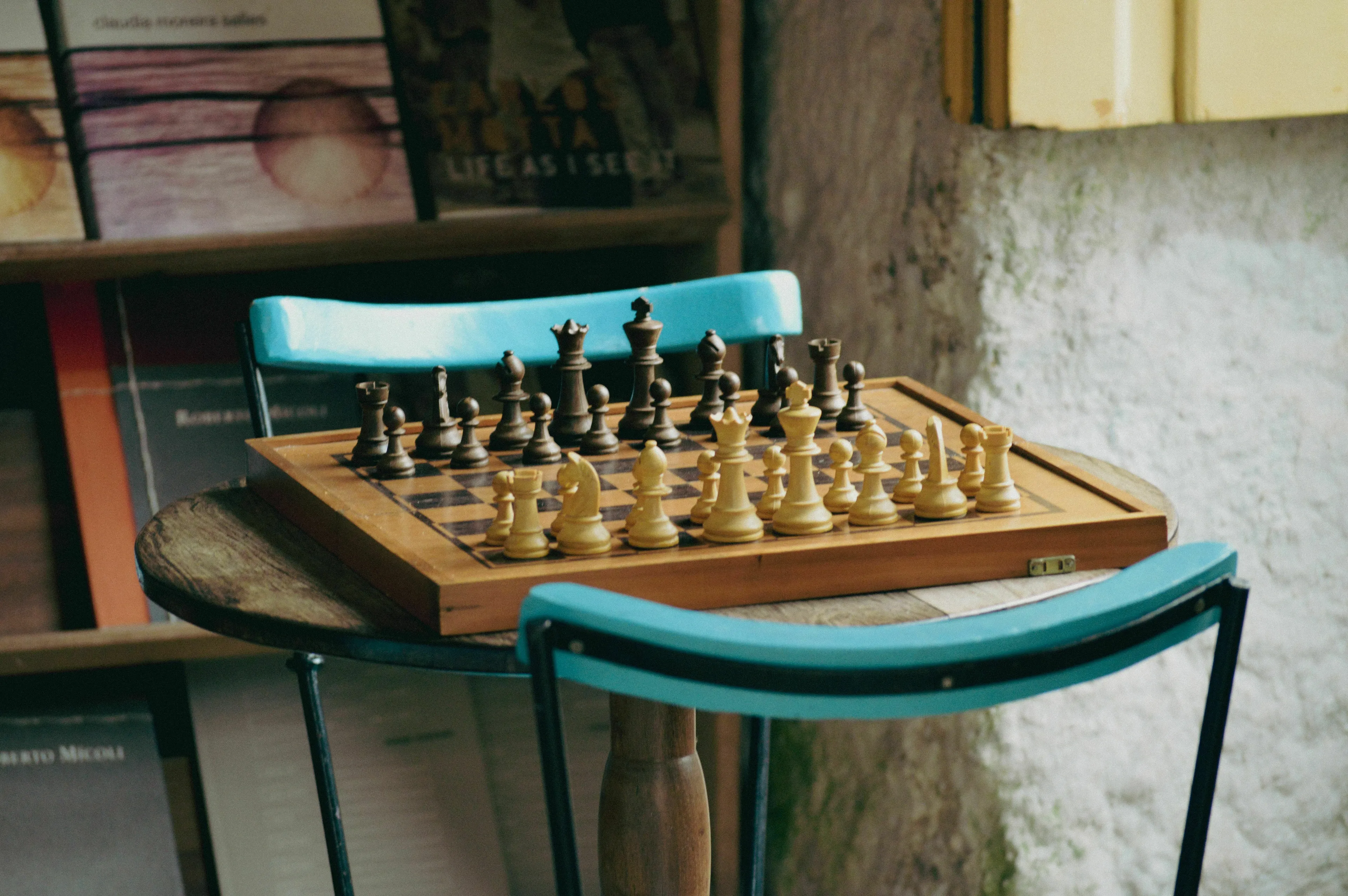 A wooden chess board with pieces set up, resting on a table between two blue chairs. This relates to general knowledge.