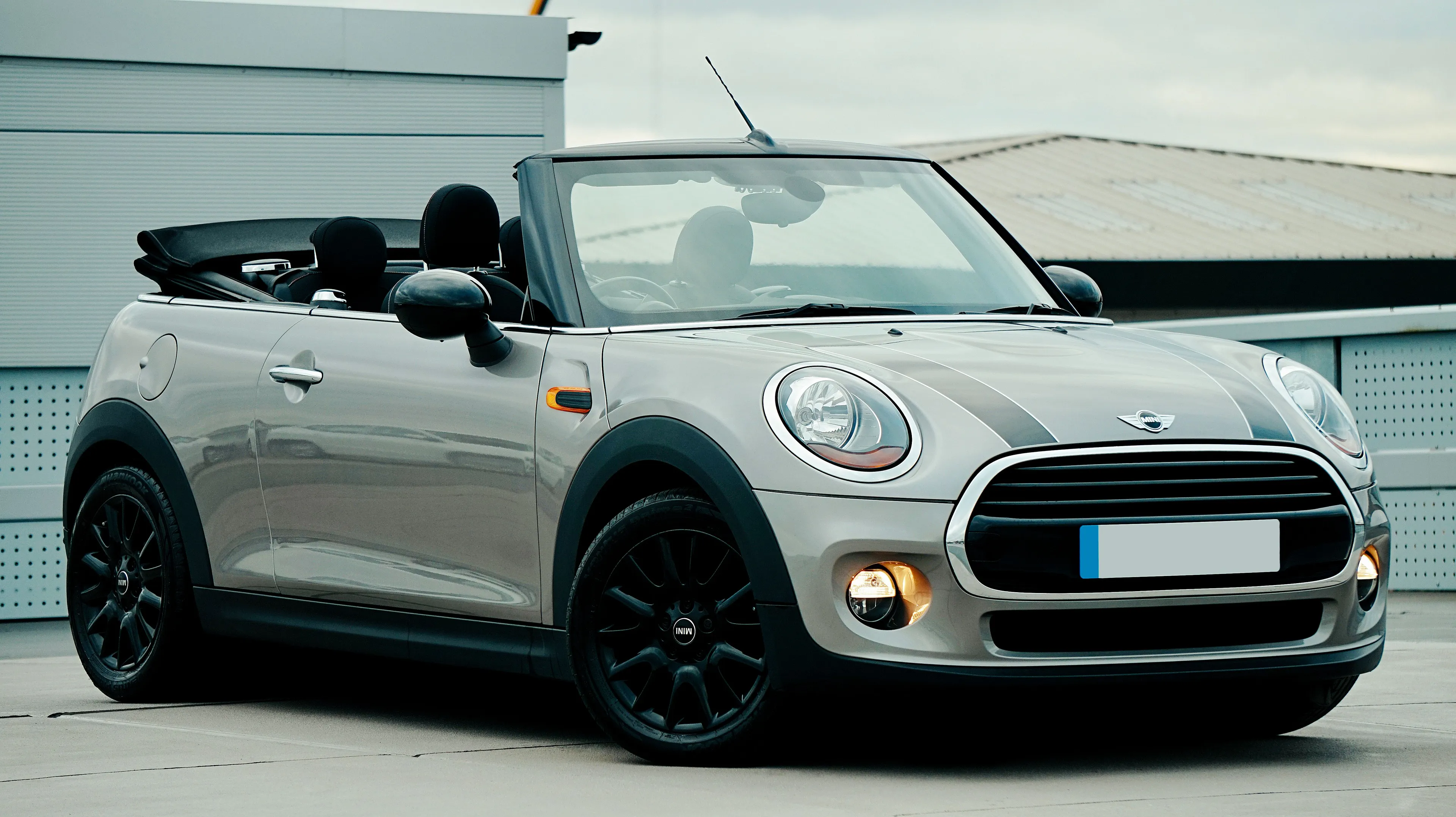 A silver Mini Cooper convertible with black stripes on the hood, parked outdoors, top down. A type of car.