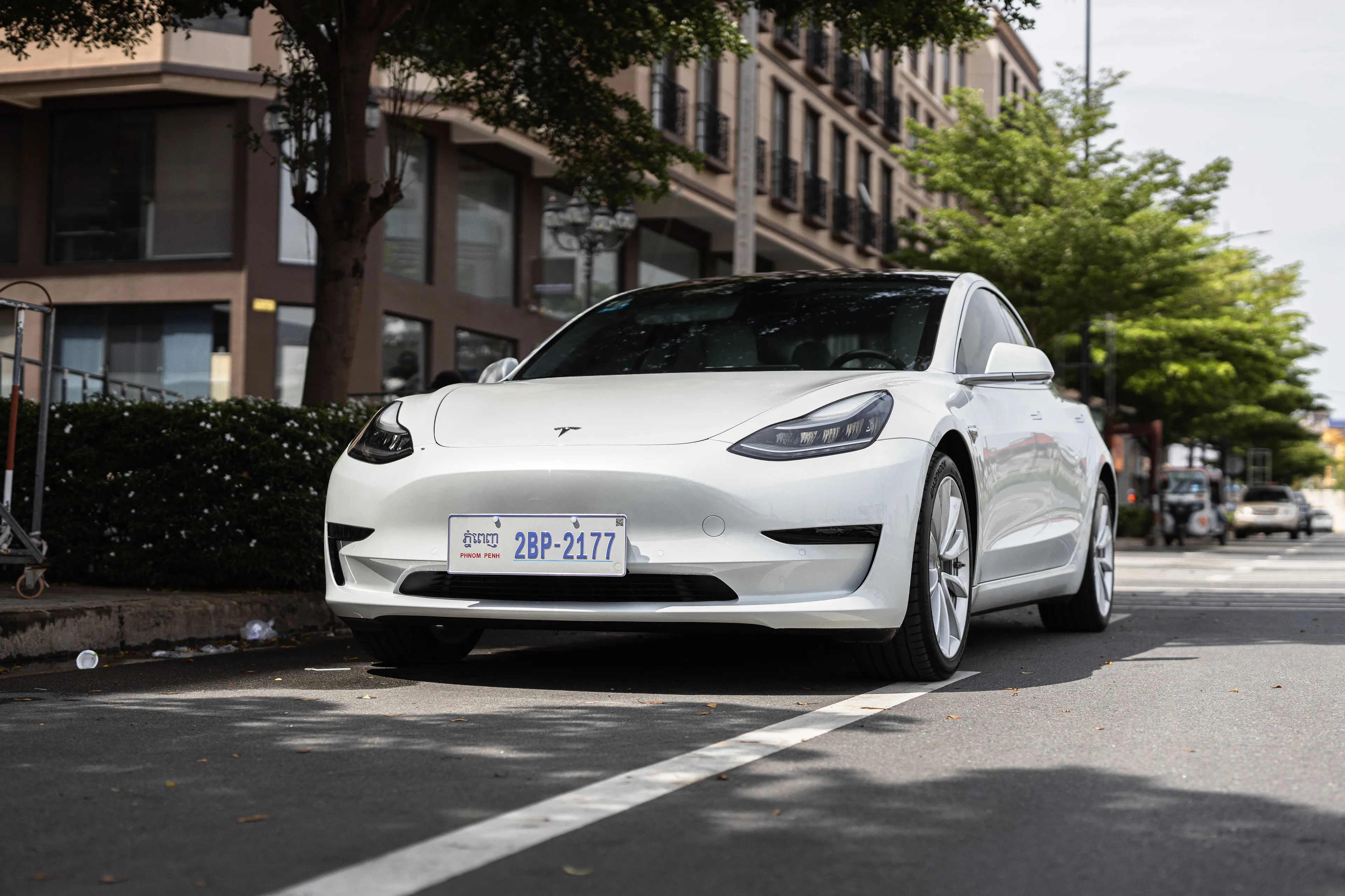 A white Tesla, a type of car, parked on a city street with buildings and trees in the background.