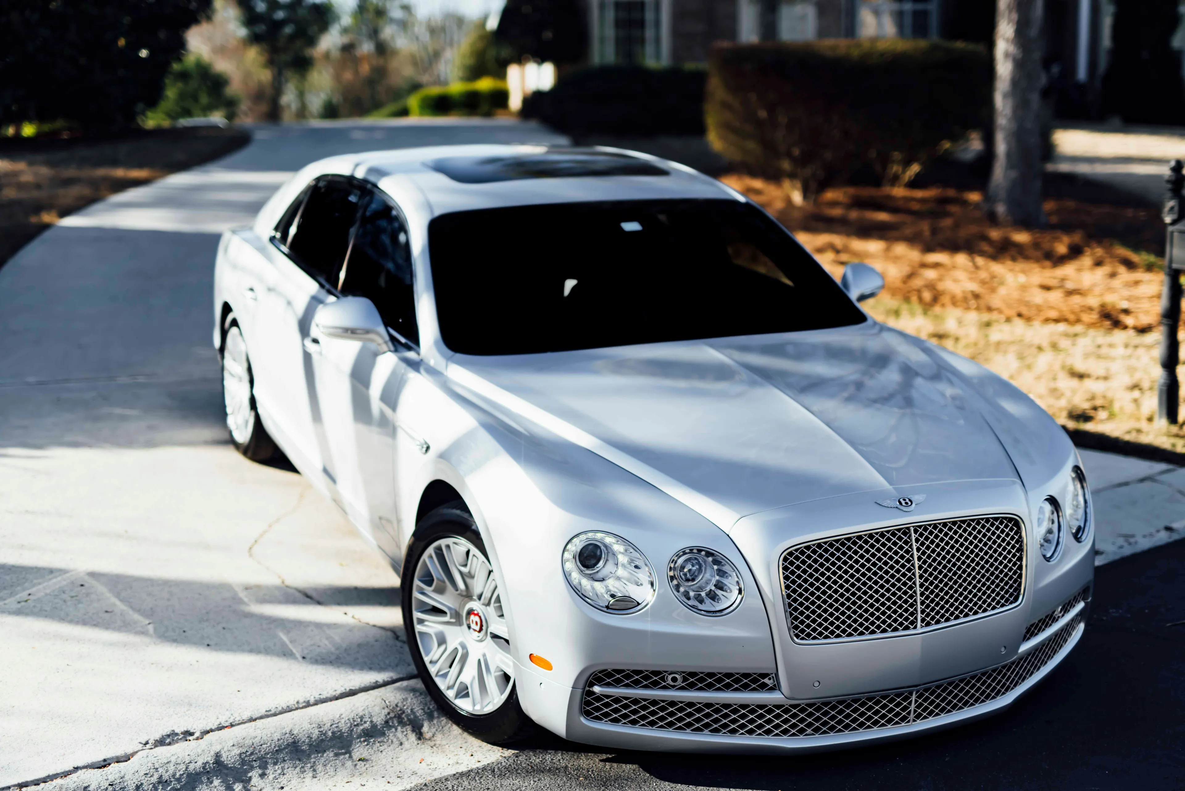 A sleek silver Bentley car, a type of car, parked on a paved driveway with a blurred background of trees and a house.