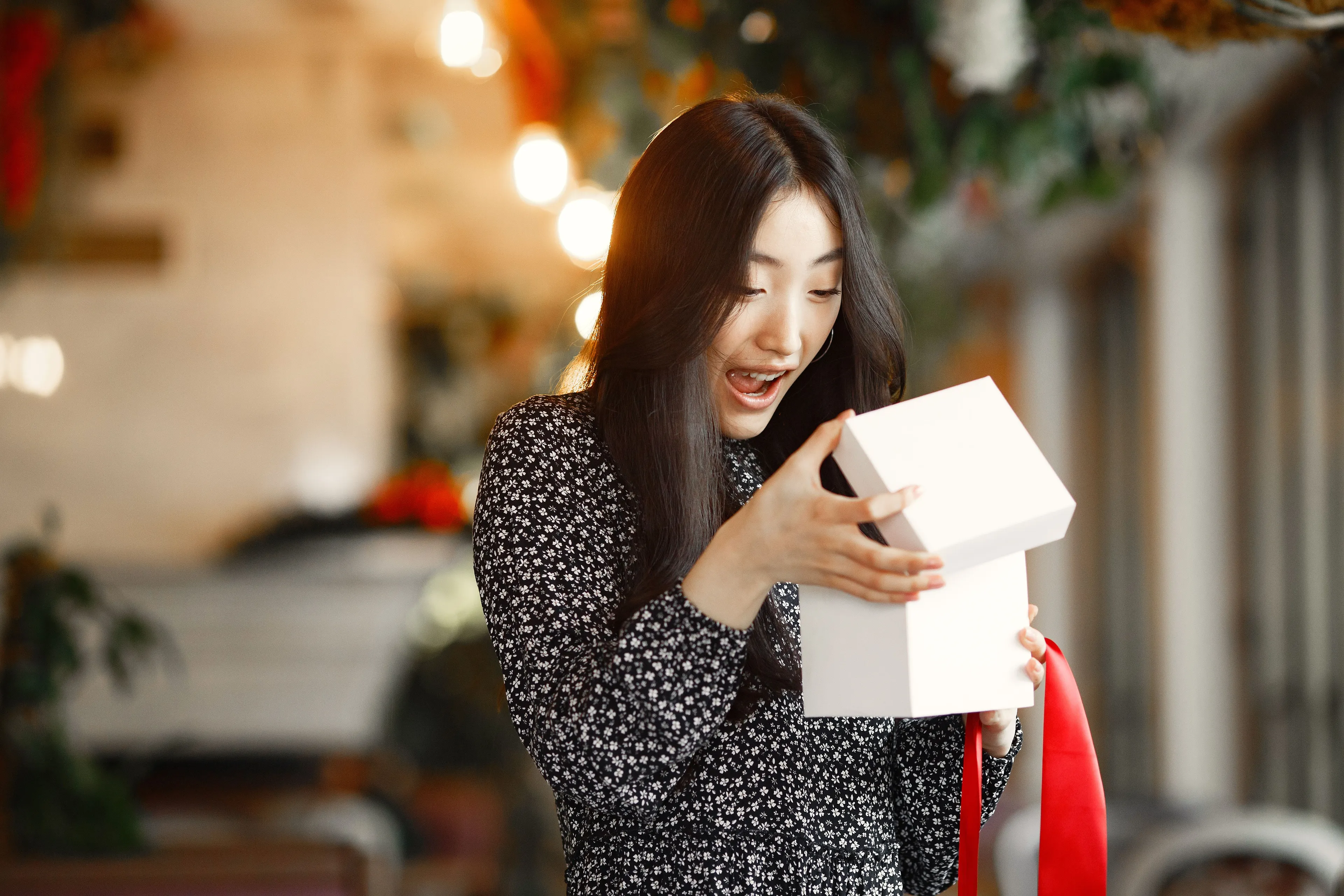 A woman with long black hair, in a floral dress, opening a gift box with a red ribbon, looking surprised. Red flag. Surprises.