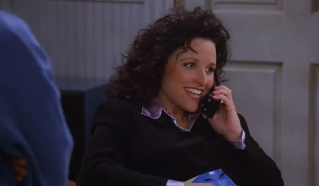Julia Louis-Dreyfus as Elaine Benes, smiling while on a flip phone. A TV catchphrase, "Yada yada yada," comes to mind.